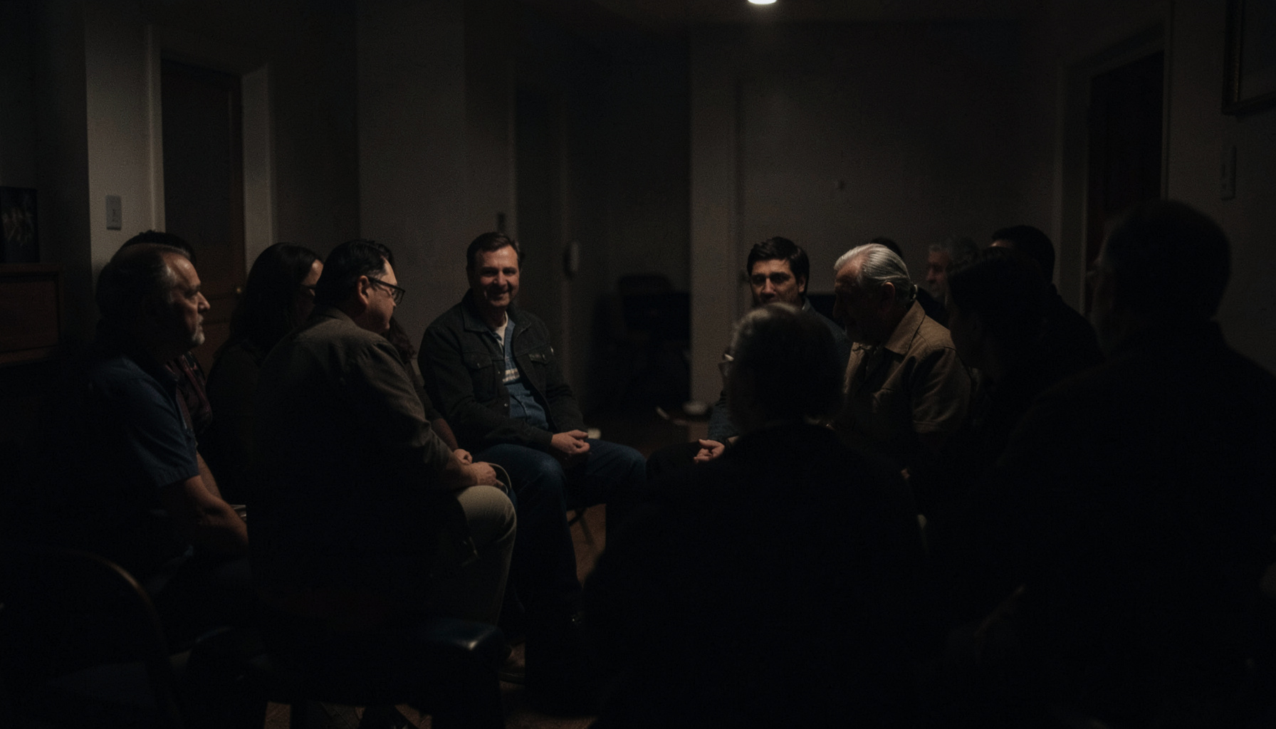 A dark, low-key photograph of a quiet support-group circle in a softly lit room, deep charcoal and muted warm tones